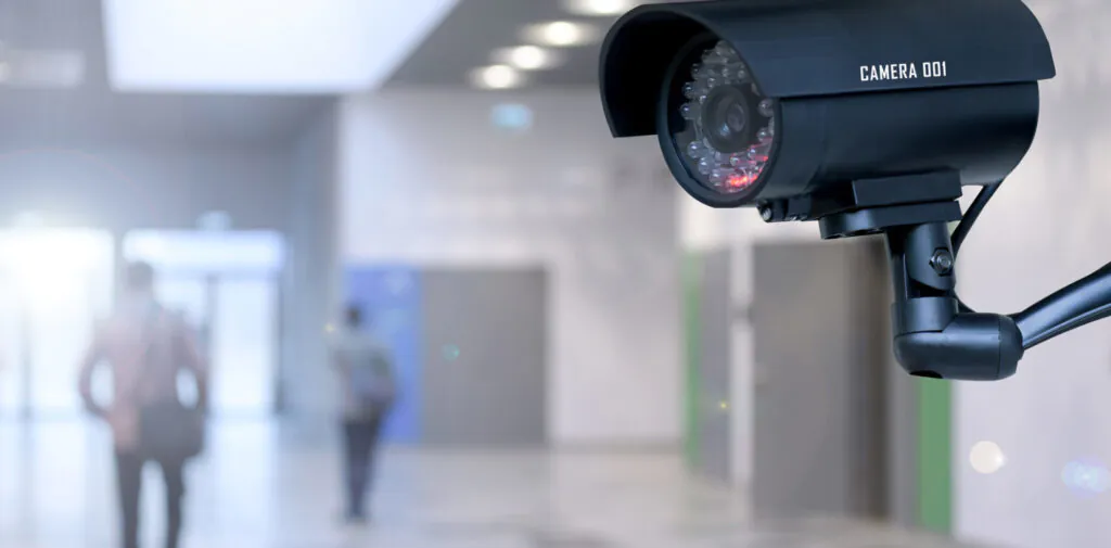 A prominent black security camera with "CAMERA 001" labeled on top is positioned in the foreground, overlooking a blurred indoor hallway with two figures walking in the distance.
