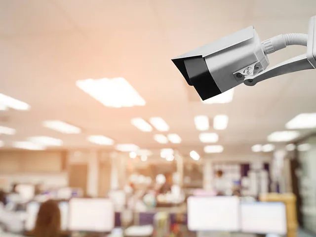 A white bullet-style CCTV camera is mounted in the upper right corner of the frame, overlooking a brightly lit, blurred office environment with cubicles and desks.