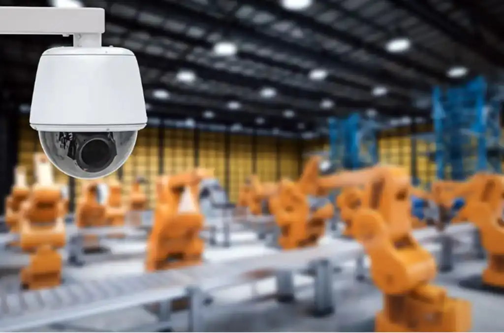 A white, spherical CCTV dome camera is mounted on a white arm in the upper left foreground, sharply in focus. Its dark lens is visible, facing towards the right. The background is a blurred yet discernible image of a large industrial facility or warehouse. Rows of orange robotic arms or machinery are visible on a conveyor system, stretching into the distance under overhead lighting. The overall impression highlights the integration of advanced CCTV monitoring and remote monitoring systems for robust intruder prevention, ensuring 24/7 monitoring and enhanced business security in an industrial or warehouse setting.
