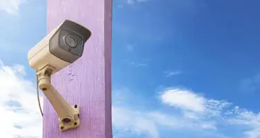 A traditional white surveillance camera is mounted on a purple post against a clear blue sky with sparse white clouds.