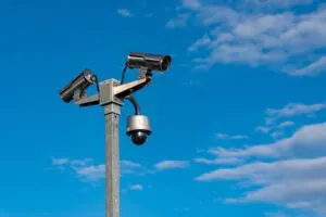 Two security cameras mounted on a pole against a clear blue sky, representing CCTV Monitoring for Business, Commercial Monitoring Service, and Live Security Monitoring. The image emphasizes the role of surveillance in ensuring Industrial Security and Workspace Security, as well as the concept of a Virtual Doorman for enhanced safety and protection.