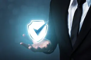 A hand in a business suit holds a glowing, stylized shield with a checkmark inside, symbolizing security and protection.