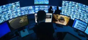 A male security supervisor points at a CCTV feed on one of many monitors while a female operator sits at a desk in a control room, demonstrating a professional monitoring center.