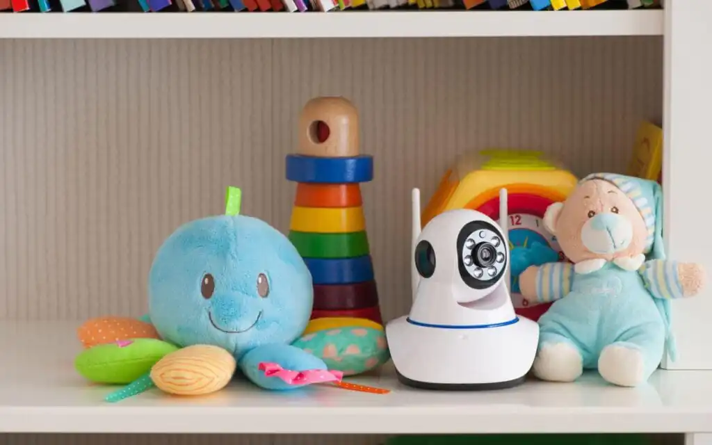 A white nanny cam with dual antennas is placed on a child’s shelf surrounded by colorful toys, including a plush blue octopus, a rainbow stacking toy, and a soft teddy bear, illustrating the use of Nanny Cams for home monitoring in a safe and child-friendly environment.
