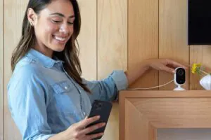 A smiling woman checks her smartphone while adjusting a small white indoor security camera on a wooden shelf, demonstrating easy access to live CCTV monitoring services at home.