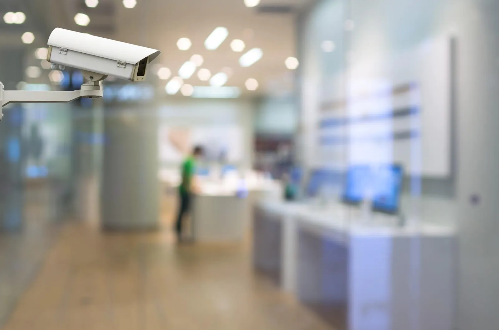 A white outdoor bullet-style CCTV camera is mounted on a wall, monitoring the entrance of a modern retail store with a customer visible inside. The store has bright, blurred lights and a glass front.