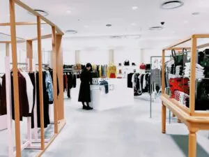 A wide-angle view of a clean, modern clothing retail store with a customer Browse merchandise. A small, black security camera is visible on the ceiling, blending into the store's design.