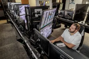 Security technician sitting at a desk with multiple computer monitors displaying real-time CCTV footage, representing 24/7 Live CCTV Monitoring Services.