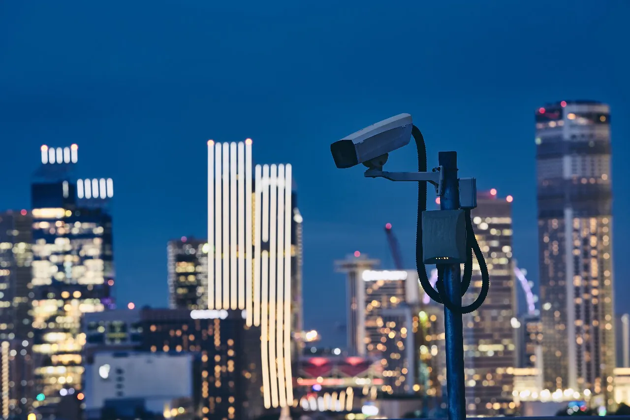 Security camera mounted on a pole at dusk with illuminated skyscrapers in the background, representing 24/7 Live CCTV Monitoring Services in the USA.
