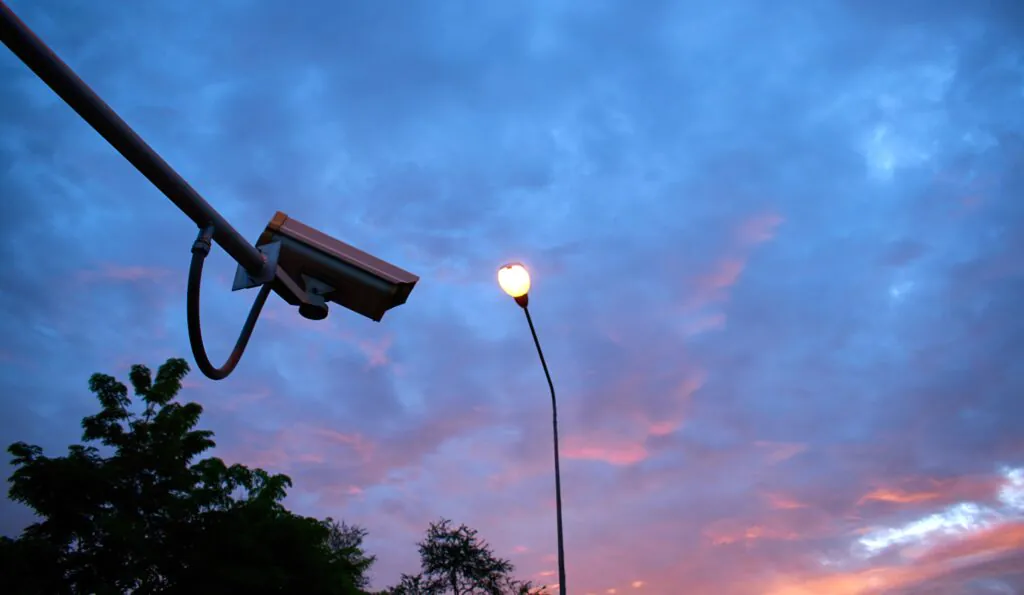 Security camera mounted on a pole next to a streetlight at dusk, representing remote monitoring services and 24/7 CCTV monitoring services.