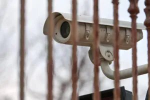 Outdoor bullet-style security camera viewed through rusted metal bars, symbolizing continuous surveillance by a trusted monitoring services provider.
