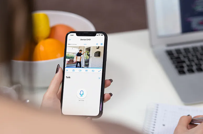 Woman holding a smartphone showing a live video feed of a delivery person at her doorstep, with a "Talk" button on the screen.