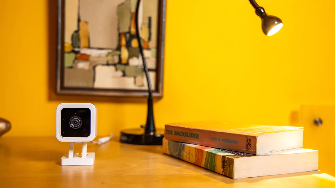 Small white square nanny camera placed on a wooden desk next to books and a lamp, against a warm yellow wall, representing stylish and effective nanny camera surveillance.