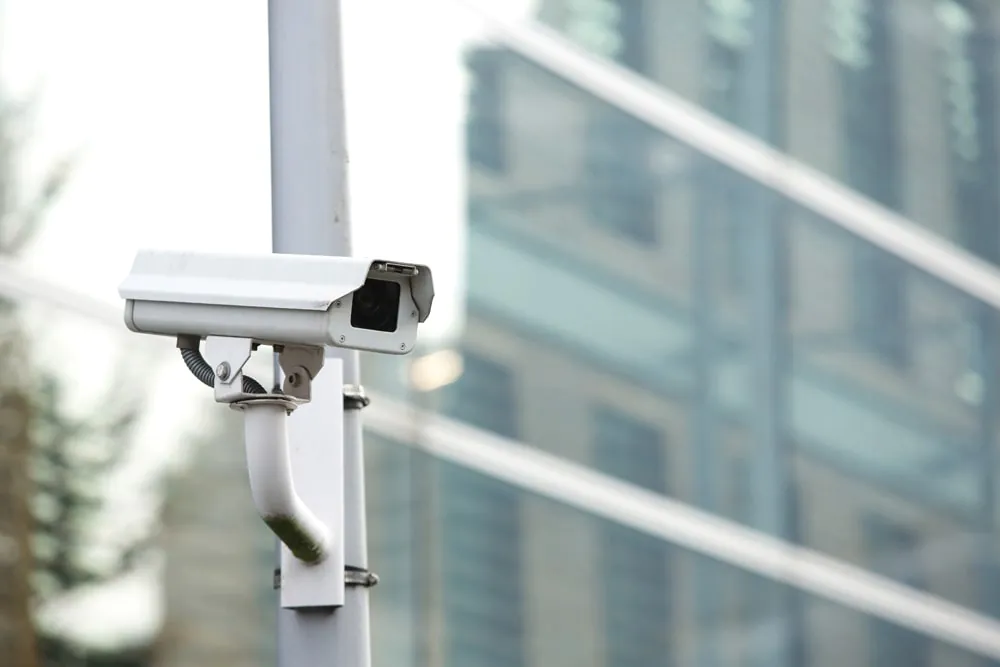 A single white outdoor security camera mounted on a pole, with a modern building in the background, representing effective CCTV Monitoring for commercial properties.