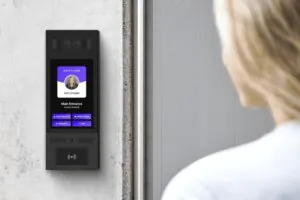A close-up view of a sleek, black digital intercom and access control panel (labeled 'Swiftlane') mounted on a wall, showing a visitor interface. A person is seen from behind approaching the device. This represents modern Virtual Doorman technology.