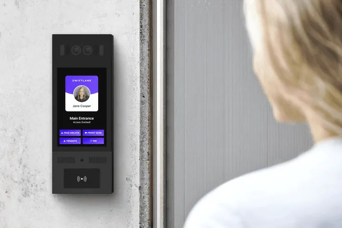 A close-up view of a sleek, black digital intercom and access control panel (labeled 'Swiftlane') mounted on a wall, showing a visitor interface. A person is seen from behind approaching the device. This represents modern Virtual Doorman technology.
