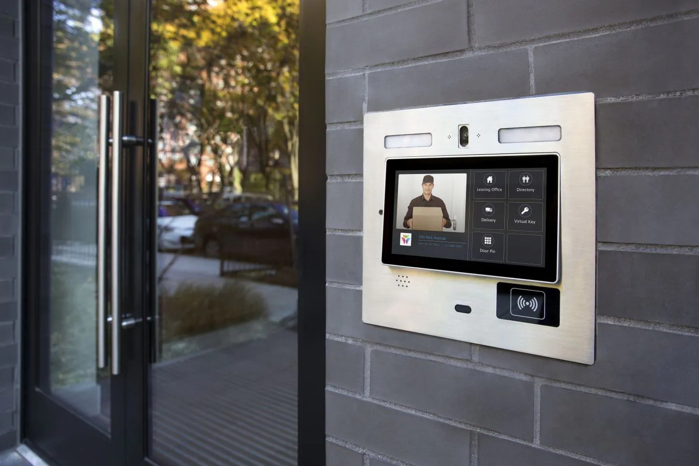 A close-up of a brushed metal intercom and display panel mounted on a brick wall next to a glass door. The screen shows a live video feed of a man holding a package, demonstrating a key function of a Virtual Doorman.