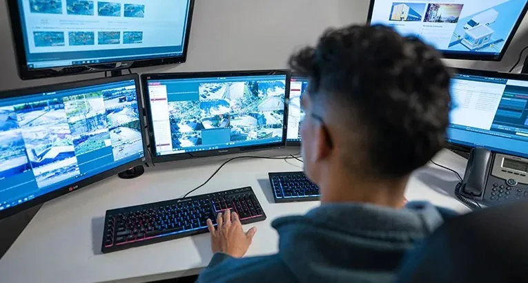 A close-up shot of an infrared security camera mounted on a textured wall in a dimly lit, blurred corridor. This type of camera is a key component of a Virtual Doorman service.