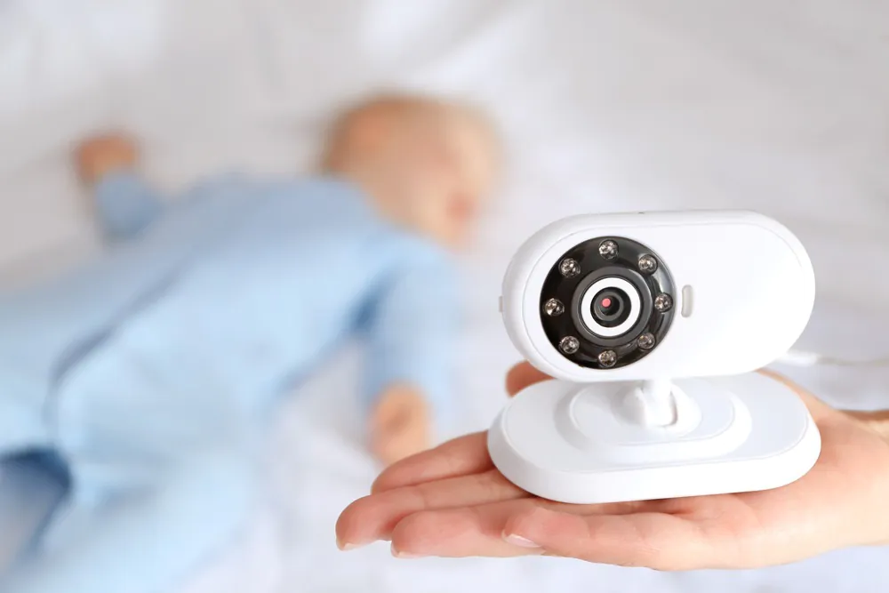 A hand holding a small, white digital baby monitor or nanny cam with infrared lights visible around the lens. In the blurred background, a baby in a blue outfit is sleeping on a white sheet.