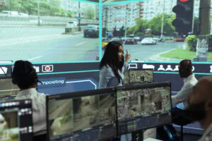A woman in a light blue shirt pointing at a large, multi-panel display showing live traffic and urban scenes, overseeing a control room with multiple operators, representing advanced CCTV surveillance.