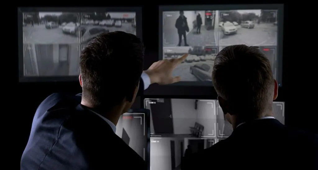 Two security professionals in suits facing multiple dark monitors displaying live CCTV feeds of an outdoor area and an indoor hallway. They are pointing to a potential security incident, emphasizing threat detection and intruder alarm through constant remote CCTV monitoring services.