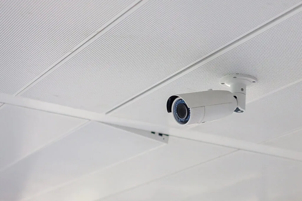 A white bullet-style security camera mounted on a suspended acoustic tile ceiling in a bright, modern interior. The installation reflects the precision of top surveillance providers.