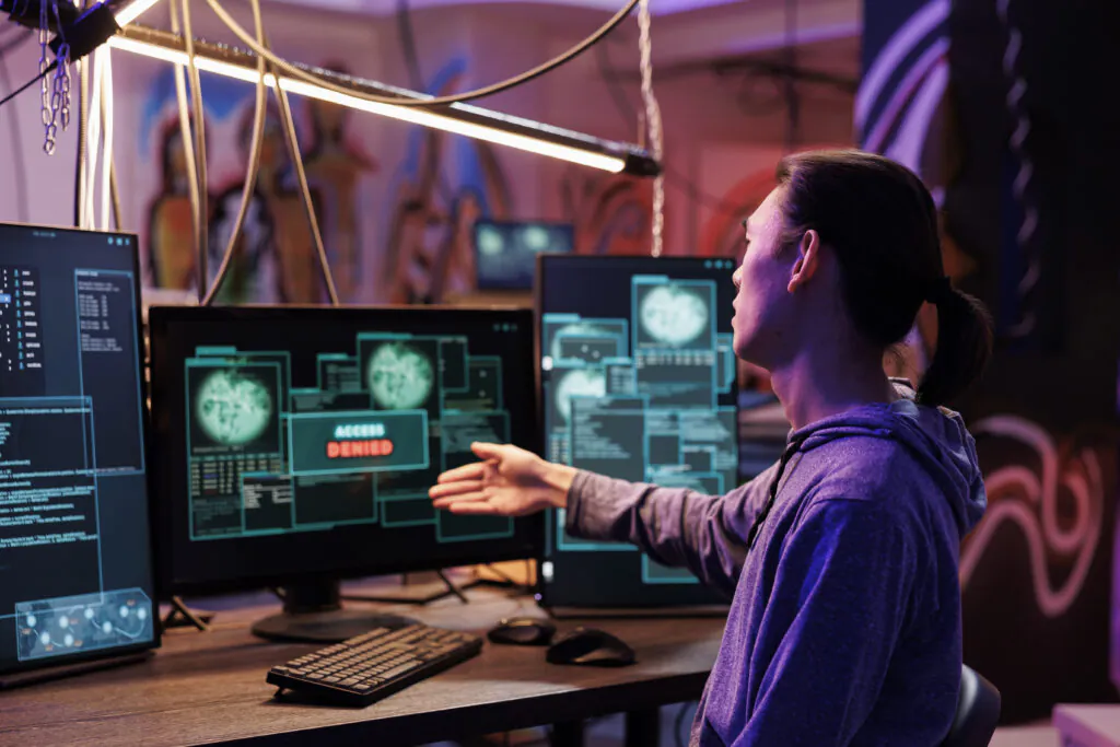A side view of a security analyst in a dark control room, gesturing toward a computer screen displaying a "ACCESS DENIED" warning along with security data and code, illustrating work in advanced Cybersecurity