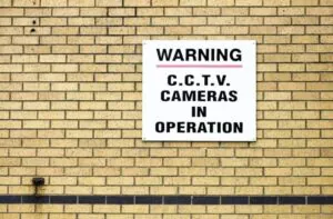 White warning sign on a brick wall reading "C.C.T.V. Cameras in Operation," confirming CCTV Surveillance.