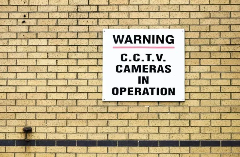 White warning sign on a brick wall reading "C.C.T.V. Cameras in Operation," confirming CCTV Surveillance.