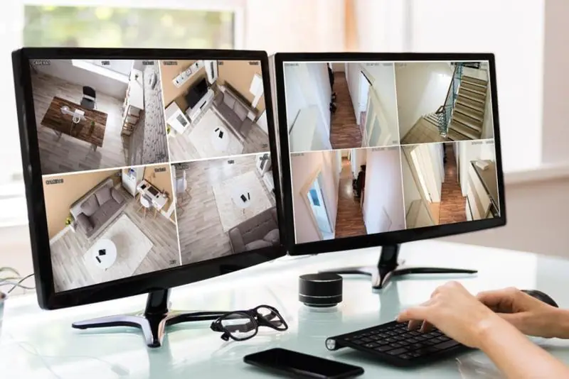 A person's hands typing on a keyboard in front of two computer screens displaying a grid of interior surveillance feeds from different rooms, highlighting the centralized service of a CCTV Monitoring Provider.
