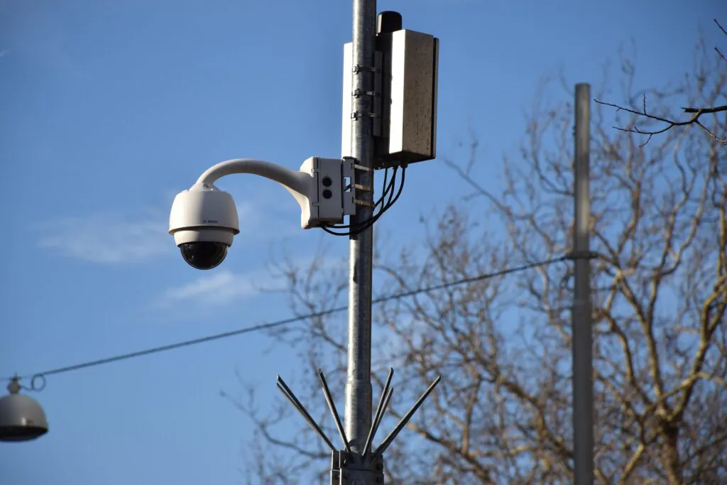 A white, adjustable outdoor CCTV camera mounted on a tall metal pole alongside a utility box, set against a blue sky, symbolizing the equipment used by a CCTV monitoring company.