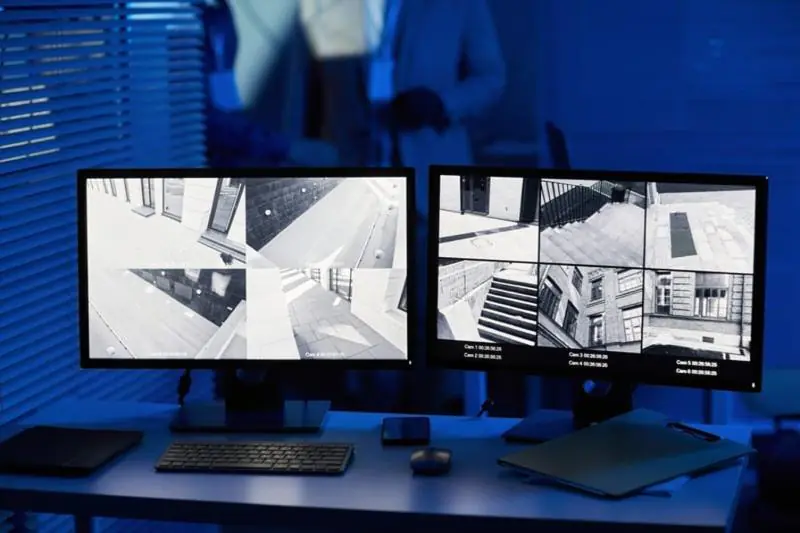Dual monitors on a desk displaying multiple black-and-white exterior camera views, signifying security monitoring services.