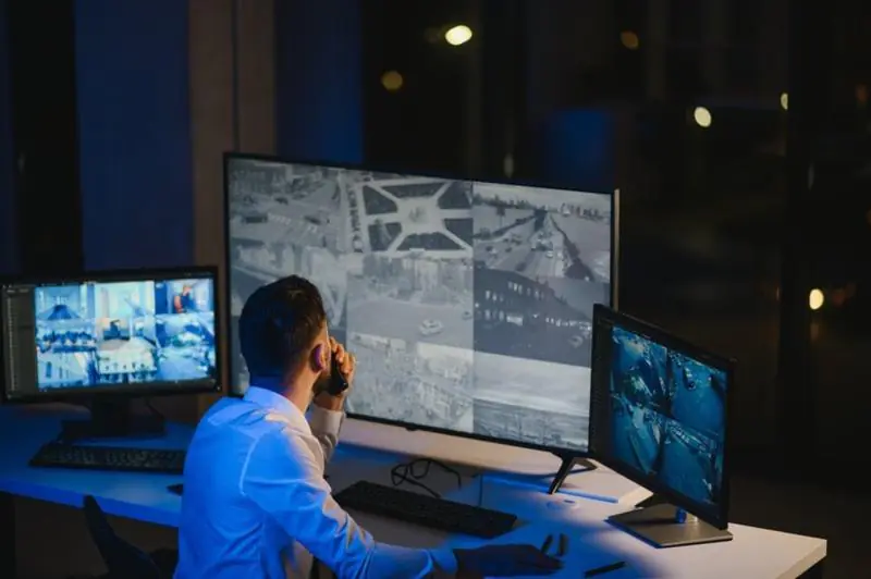 A security professional sitting in a dark control room at night, talking on a phone while monitoring several large screens showing various black-and-white CCTV Surveillance feeds of traffic and streets.