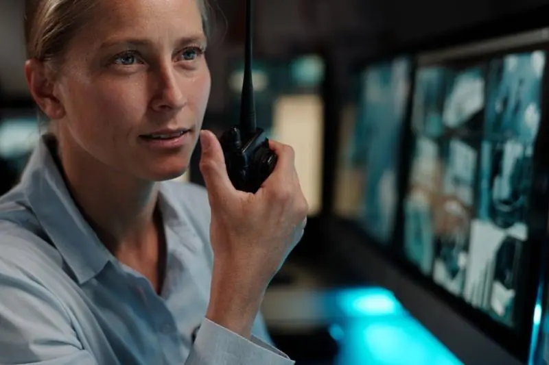 A female security monitor operator sitting in a dark room, speaking into a walkie-talkie while monitoring a large screen split into four interior surveillance feeds, showing active surveillance services.