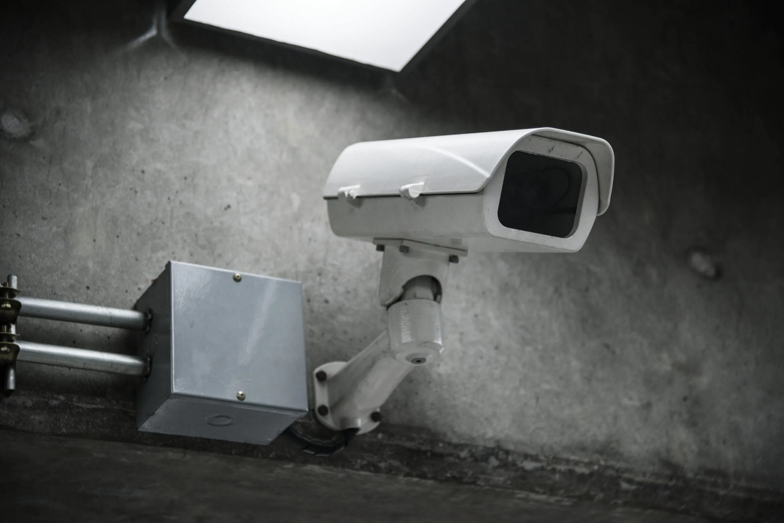 A white, outdoor-style CCTV camera mounted on a textured gray concrete wall, connected to a metal junction box, representing the equipment used for professional CCTV monitoring.