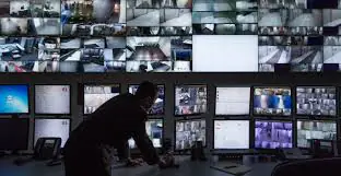 A person standing in front of a wall of screens displaying multiple video feeds, likely in a CCTV monitoring center.