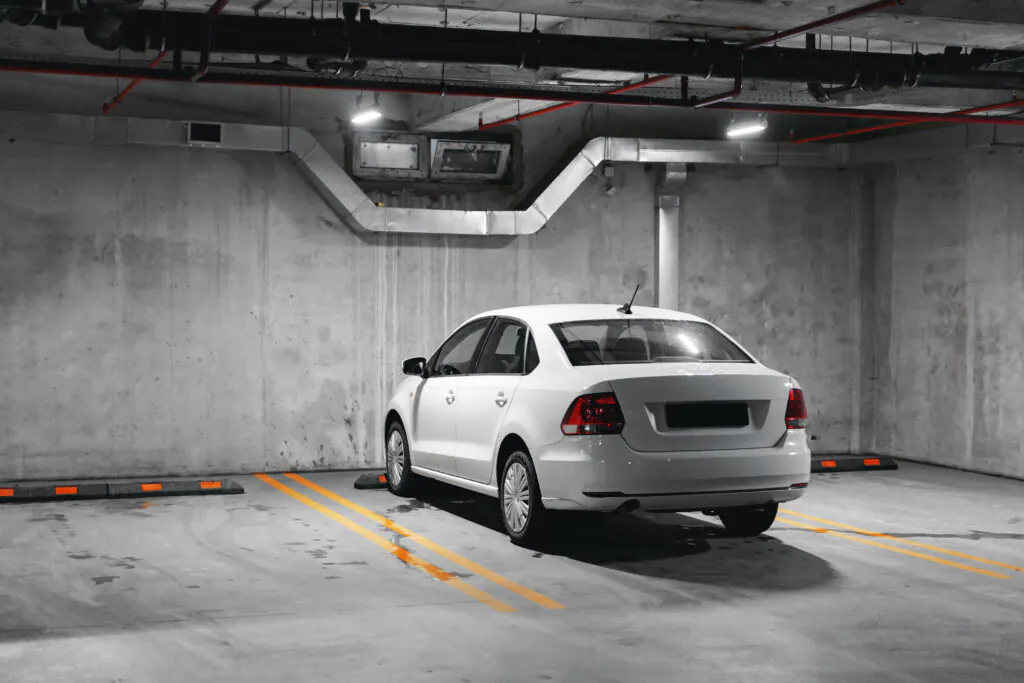 A single white sedan parked in a designated yellow-lined space against a textured concrete wall in a clean, brightly lit underground parking structure, highlighting the need for isolated-spot Parking Lot Surveillance.