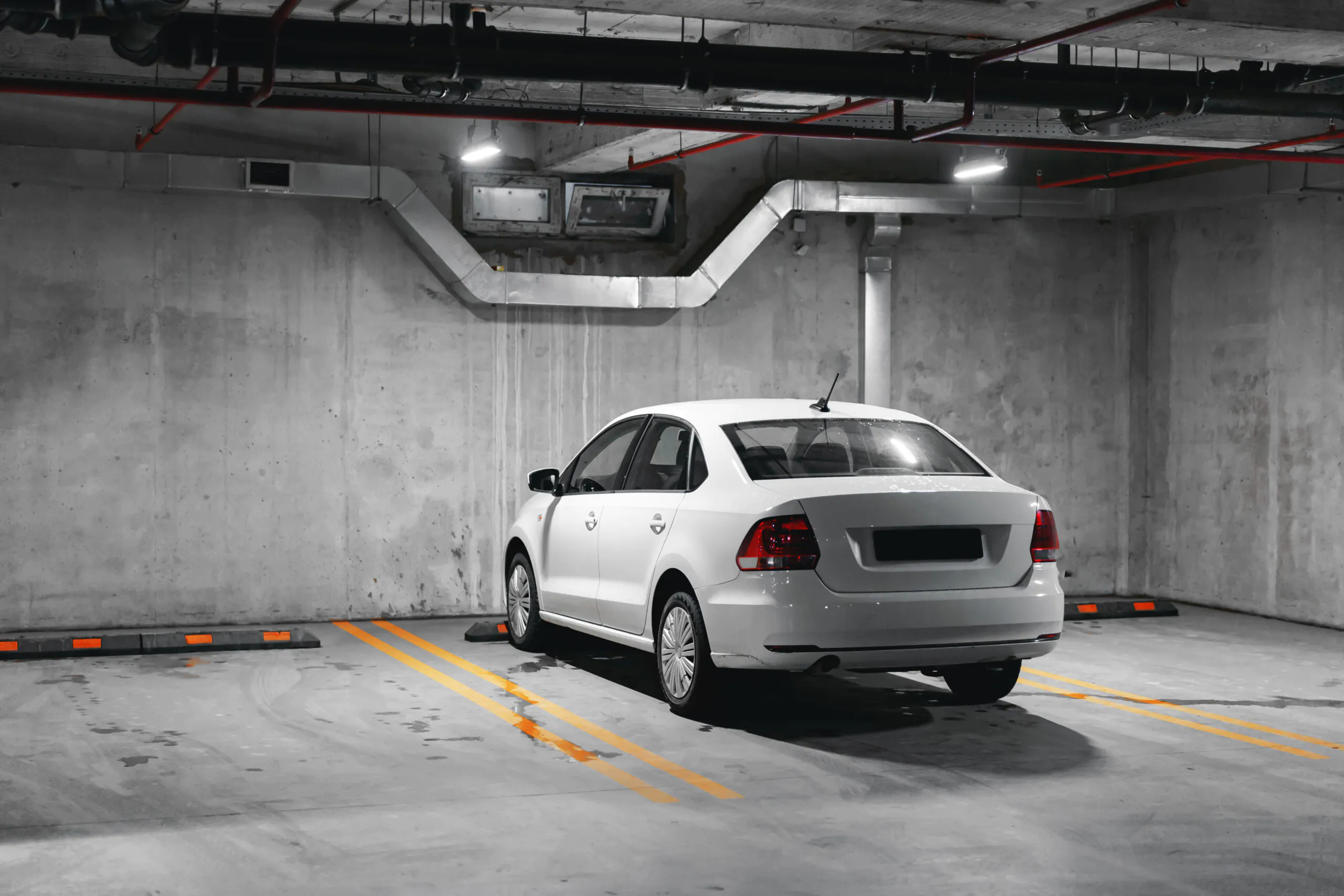 A single white sedan parked in a designated yellow-lined space against a textured concrete wall in a clean, brightly lit underground parking structure, highlighting the need for isolated-spot Parking Lot Surveillance.