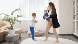 A mother holding a baby while a young boy stands nearby, all standing barefoot on a circular rug in a brightly lit living room with large windows, representing a safe family environment supported by Nanny Cams Monitoring.