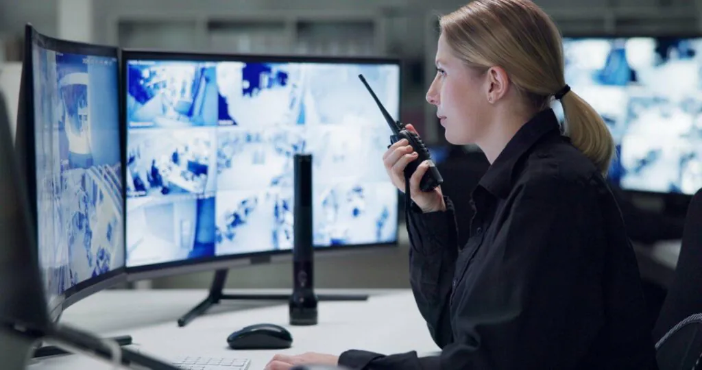 Female security officer with a ponytail speaking on a radio next to monitors, key for two-way audio surveillance.
