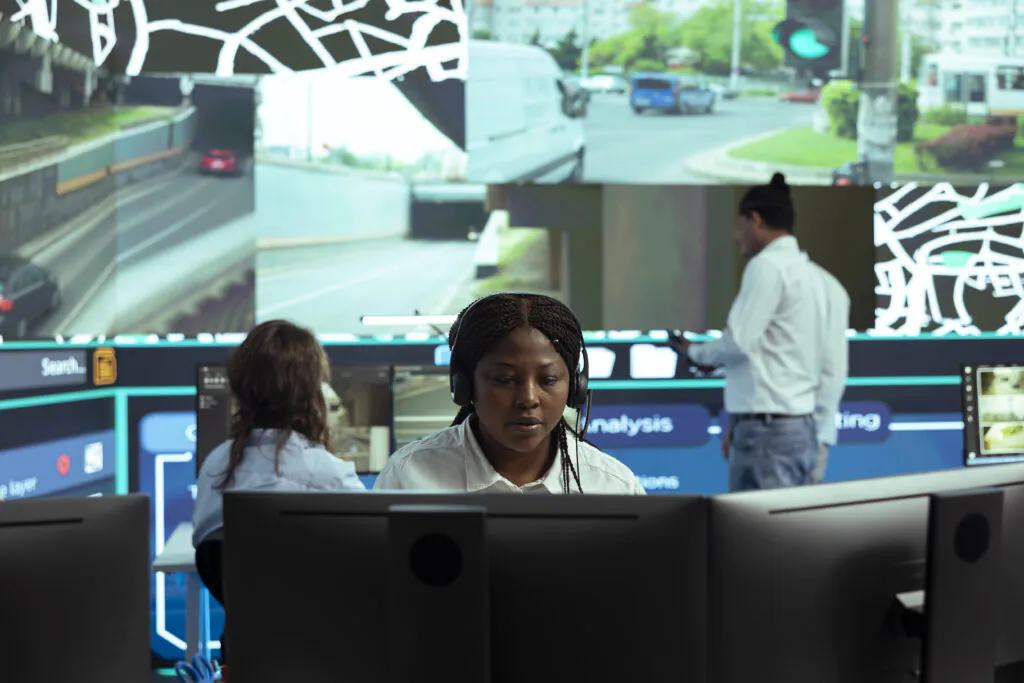 A female operator wearing a headset monitors a massive video wall for remote CCTV surveillance in a control room.