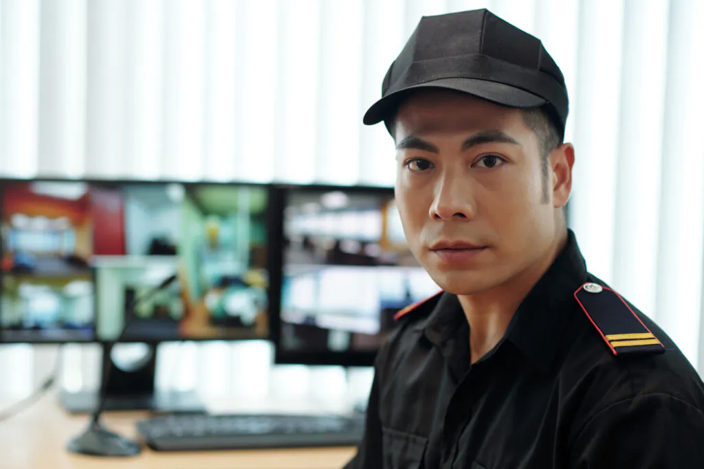 A close-up portrait of a security guard in a black uniform cap sitting in front of monitoring screens, representing professional remote CCTV services