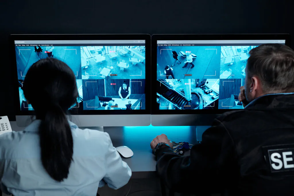 Two security professionals, one with a "Security" patch, viewing blue-tinted screens to provide remote CCTV monitoring for small business security
