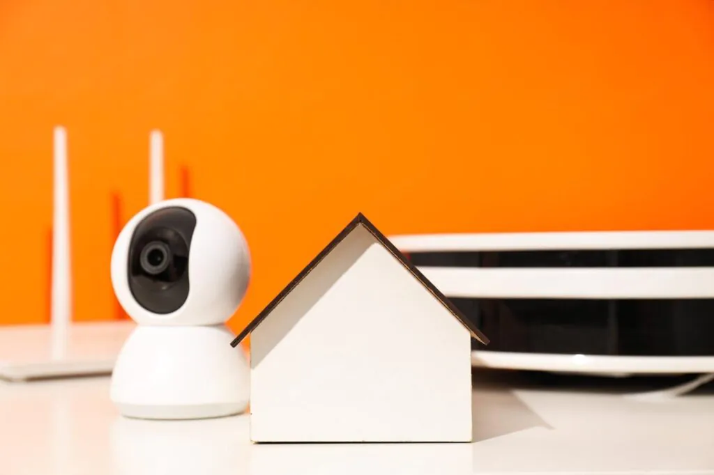 A small model house sits next to a wireless camera and router against an orange background, representing Video Surveillance for the home
