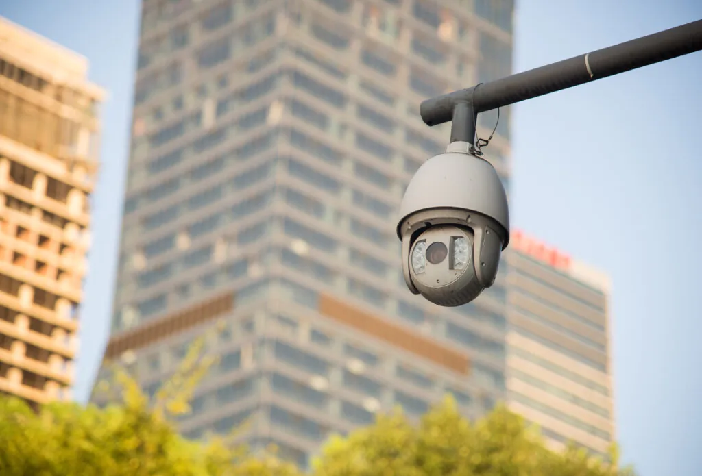 A white PTZ dome camera mounted on a metal arm overlooking an urban landscape, illustrating the role of surveillance and CCTV surveillance in city safety