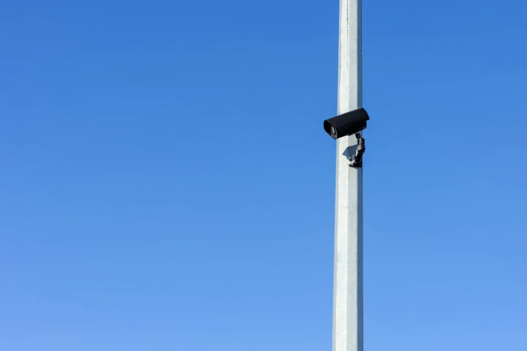 A black security camera mounted high on a metal pole against a clear blue sky, illustrating the infrastructure needed for effective CCTV surveillance and crime prevention