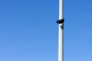A black security camera mounted high on a metal pole against a clear blue sky, illustrating the infrastructure needed for effective CCTV surveillance and crime prevention