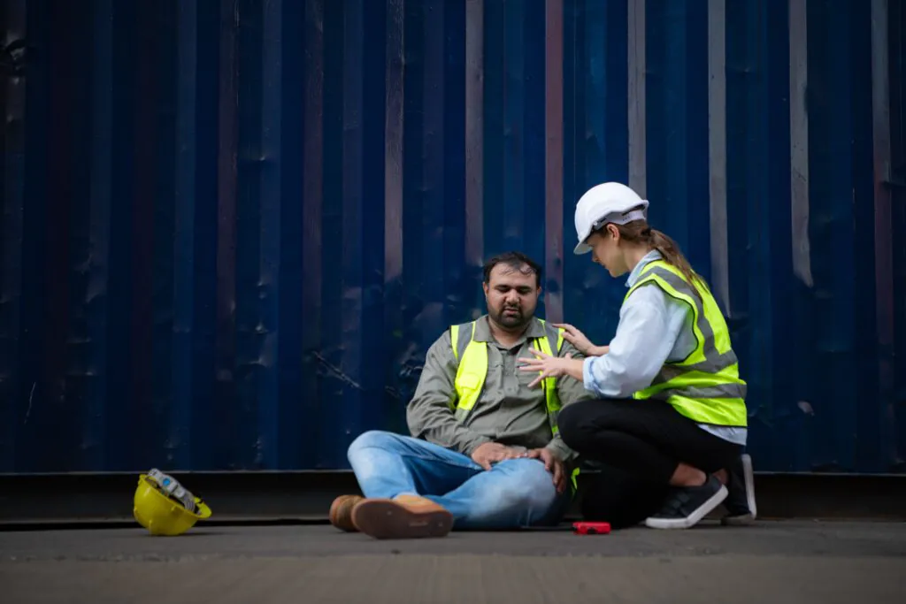 A female construction worker aiding an injured colleague near a blue shipping container, a scenario where remote CCTV monitoring ensures quick incident response