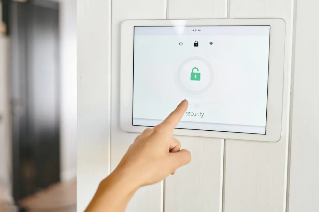 A person's hand using a touchscreen control panel to activate security monitoring, displaying a green lock icon for residential security. Security Monitoring Company