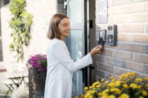 A woman in a white blazer smiling while using a keycard on a digital intercom system at the entrance of a building, representing the convenience of modern Virtual Doorman Services